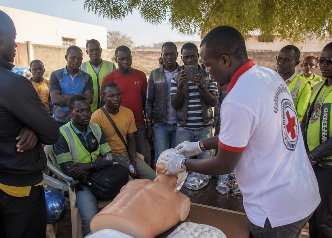 Transport drivers gathered outdoors watching an instructor demonstrate resuscitation on a training mannequin
